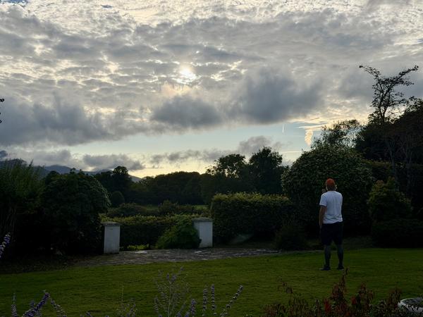 A lone figure stands in a lush green garden at The Fossa Way, gazing out at a panoramic view of distant mountains and dense forests under a dramatic, cloudy sky